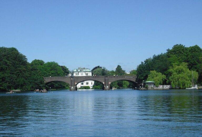 Klassenfahrt Hamburg Kanutour Alster. Während der Klassenfahrt nach Hamburg unternimmt die Schulklasse eine Kanutour über die Alster und die Kanäle. Auf dem Bild sehen wir eine alte Alsterbrücke welche zwei grüne Ufer verbindet. Im Hintergrund ein weißes, mondänes Gebäude. Auf dem Wasser einige Paddler.